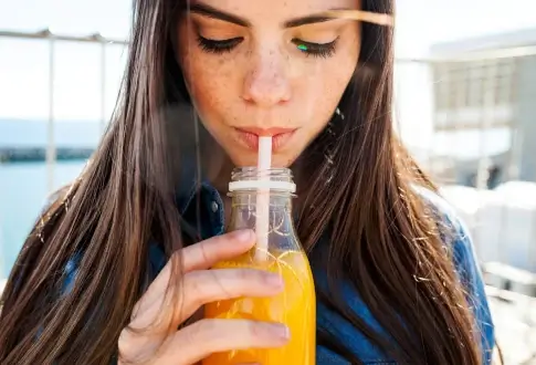 На фото красивая девушка с веснушками и длинными волосами пьёт апельсиновый сок через трубочку