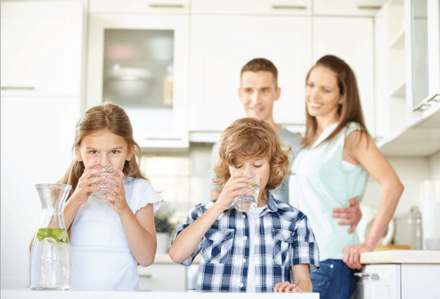 На фото счастливая семья, заказавшая бутилированную воду Aqua Plus с двумя детьми. Дети с наслаждением пьют воду