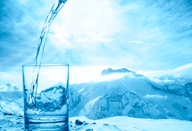 На фото в прозрачный стеклянный стакан наливают кристально чистую водую На заднем фоне- заснеженные горы
